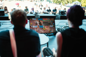 Mario Kart Turnier-Finale Zuschauer genießen ein aufregendes Videospielturnier während des Elbenwald Festivals. Auf dem Bildschirm sind vier Spieler in einem Rennspiel zu sehen, während die Menge im Hintergrund gespannt zuschaut.
