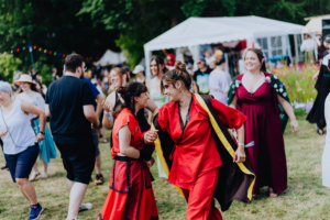 Historische Tänze Zwei Frauen in roten Kostümen tanzen fröhlich auf dem Elbenwald Festival, umgeben von einer bunten Menschenmenge. Im Hintergrund sind Zelte und festliche Lichter sichtbar, die die lebendige Atmosphäre des Festivals unterstreichen.