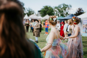Historische Tänze Festivalbesucherinnen in bunten Kostümen tanzen auf dem Elbenwald Festival, umgeben von Ständen und anderen Teilnehmern. Die fröhliche Atmosphäre und kreative Outfits spiegeln das nerdige Flair des Events wider.