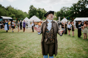 Historische Tänze Ein Mann in historischer Kleidung, der auf dem Elbenwald Festival spricht, umgeben von Besuchern in verschiedenen Kostümen. Im Hintergrund sind Festzelte und eine lebendige Festivalatmosphäre zu sehen, die die Verbindung von Musik und Nerd-Kultur zeigt.