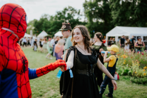 Historische Tänze Ein rot-blauer Spider-Man und eine lächelnde Frau in einem schwarzen Kleid halten sich bei der Elbenwald Festival zusammen an der Hand. Im Hintergrund sind weitere Festivalbesucher und Zelte zu sehen, die die lebendige Atmosphäre des Events unterstreichen.