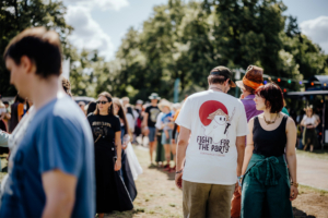 Historische Tänze Menschen auf dem Elbenwald Festival, das Musik und Nerd-Kultur vereint. Im Vordergrund ein Mann mit T-Shirt und eine Frau in lässiger Kleidung, umgeben von einer bunten Menge und festlicher Atmosphäre im Freien.