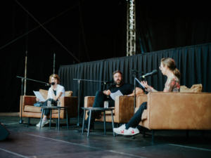 True Lausitz Drei Sprecher sitzen auf einer Bühne beim Elbenwald Festival, umgeben von Mikrofonen und Notizen. Die Atmosphäre ist entspannt, während sie sich auf eine Diskussion oder Lesung vorbereiten. Im Hintergrund sind die Bühnenvorbereitungen sichtbar.