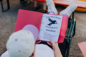 Ein Teilnehmer des Elbenwald Festivals hält ein pinkes Liederbuch mit dem Titel "Camp Half-Blood SongBook" in den Händen. Das Buch enthält Informationen zu Mitsingen und Verhaltensregeln für die musikalische Unterhaltung auf dem Festival.