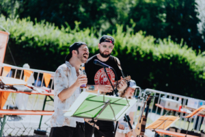 Ukulele spielen mit Tag der Helden Zwei Musiker treten auf dem Elbenwald Festival auf, einer singt begeistert und hält eine Getränkeflasche, während der andere auf einer Ukulele spielt. Im Hintergrund sind Ständer für Notenblätter und Festivalbänke sichtbar, umgeben von grüner Natur.
