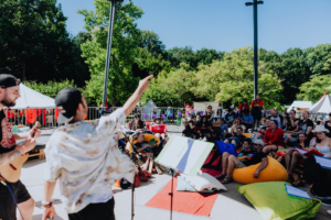 Ukulele spielen mit Tag der Helden Musiker führen auf der Bühne beim Elbenwald Festival auf, während das Publikum auf bunten Sitzkissen entspannt. Die Atmosphäre ist lebhaft und fröhlich, umgeben von Bäumen und Festivalzelten. Ein perfekter Tag für Musik und Gemeinschaft.