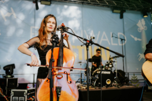 Vogelsang Eine Musikerin spielt Cello auf der Bühne des Elbenwald Festivals, umgeben von Mikrofonen und Instrumenten. Im Hintergrund ist ein Schlagzeuger zu sehen, während die Festival-Atmosphäre mit bunten Lichtern und einer lebhaften Kulisse ergänzt wird.