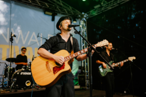 Vogelsang Ein Musiker spielt Gitarre auf der Bühne des Elbenwald Festivals, während ein Schlagzeuger im Hintergrund die Band unterstützt. Die Atmosphäre ist lebhaft und farbenfroh, ideal für Fans von Musik und Nerd-Kultur.
