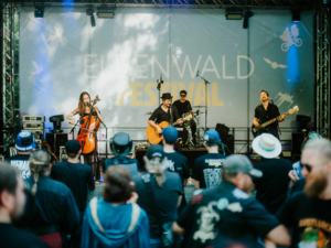 Vogelsang Auf der Bühne des Elbenwald Festivals spielt eine Band mit Cello, Gitarre und Bass. Im Vordergrund stehen zahlreiche Zuschauer in kreativen Outfits, die die Musik genießen. Die Atmosphäre ist lebhaft und feierlich, umgeben von Bäumen und Festival-Elementen.