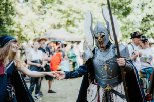 Ein Teilnehmer des Elbenwald Festivals in beeindruckender Rüstung mit Flügeln schüttelt die Hand einer Frau in einem historischen Kostüm. Im Hintergrund sind weitere Festivalbesucher und bunte Kostüme zu sehen, die die lebendige Atmosphäre des Events widerspiegeln.