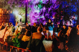Werwolf-Runde zur Nacht Eine Gruppe von Festivalbesuchern sitzt im Halbrund und hört einer Rednerin zu, während bunte Lichter die Umgebung erhellen. Die Szene zeigt die entspannte Atmosphäre des Elbenwald Festivals, wo Musik und Gemeinschaft im Vordergrund stehen.