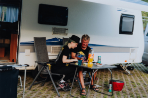 Zwei Festivalbesucher sitzen lächelnd an einem Tisch vor einem Wohnmobil, während sie gemeinsam Essen zubereiten. Die entspannte Atmosphäre und die Festivalbekleidung unterstreichen die Vorfreude auf das Elbenwald Festival.