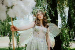 Eine fantasievolle Frau mit Elfenflügeln und Blumenkranz hält einen dekorativen Sonnenschirm. Sie steht vor einem grünen Hintergrund beim Elbenwald Festival, das Musik und Convention-Elemente kombiniert und eine kreative Gemeinschaft feiert.
