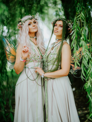 Zwei Frauen in fantasievollen Elbenkostümen mit Flügeln und Blumenkränzen posieren in einer grünen Umgebung beim Elbenwald Festival. Die kreative Atmosphäre und die detailreiche Kostümierung spiegeln den zauberhaften Geist des Festivals wider.