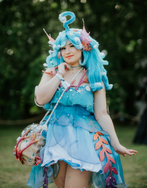 Eine junge Frau mit blauen Haaren und einem fantasievollen Kostüm aus transparentem Stoff posiert beim Elbenwald Festival. Ihr Outfit ist mit Muscheln und Seesternen verziert, während sie eine kreative Tasche mit Meeresmotiven trägt.