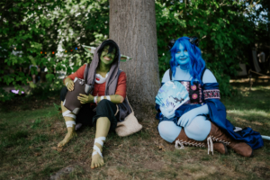 Zwei Cosplayer sitzen unter einem Baum beim Elbenwald Festival. Die Person links hat grüne Haut und trägt ein rotes Oberteil, während die Person rechts blaue Haut hat und einen Fächer hält. Die Umgebung ist grün und freundlich, ideal für ein Festival.