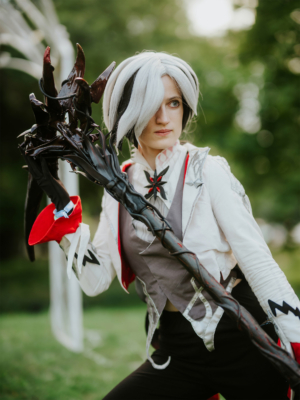 Cosplayer mit silbernem Haar und schwarzem Stab auf dem Elbenwald Festival. Die Person trägt ein auffälliges Kostüm mit roten und grauen Elementen, während sie in einer dynamischen Pose steht und die grüne Umgebung im Hintergrund betont.