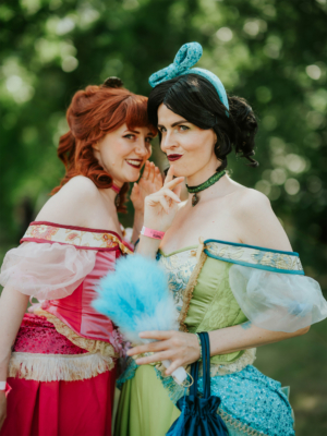 Zwei Frauen in bunten, fantasievollen Kostümen posieren fröhlich auf dem Elbenwald Festival. Die eine trägt ein pinkes Kleid mit Rüschen, während die andere in einem grünen Kleid mit blauen Akzenten strahlt. Im Hintergrund ist eine grüne, sonnige Umgebung zu sehen.