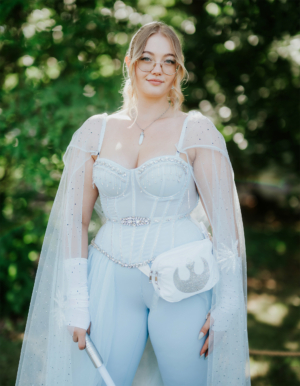 Eine Cosplayerin in einem eleganten, hellblauen Outfit mit funkelnden Akzenten und einem transparenten Umhang, steht im Freien. Der Hintergrund zeigt eine grüne, natürliche Umgebung, passend zum Elbenwald Festival, das Musik und Nerd-Kultur vereint.