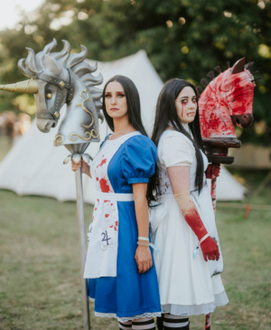 Zwei Cosplayerinnen stehen Rücken an Rücken auf dem Elbenwald Festival. Die erste trägt ein blutverschmiertes blaues Kleid, während die zweite in einem weißen Kleid mit blutigen Akzenten gekleidet ist. Beide halten stilisierte Pferde-Stäbe in der Hand.