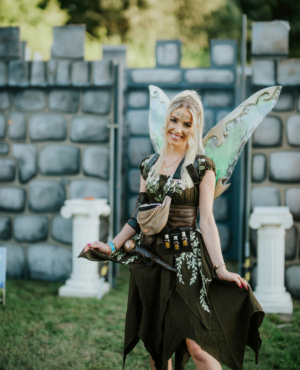Ein fröhliches Festivalbesucher-Mädchen mit Feenflügeln und fantasievoller Kleidung posiert vor einer steinernen Kulisse auf dem Elbenwald Festival, umgeben von grüner Wiese und dekorativen Säulen. Ideal für Fans von Fantasy und Musik.