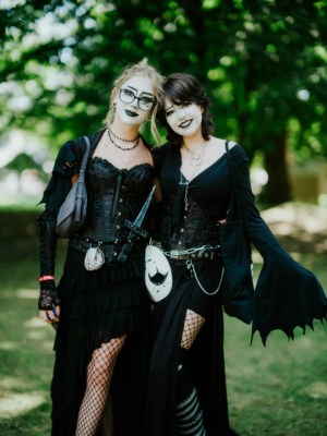 Zwei Frauen in schwarzer Gothic-Kleidung mit auffälligem Make-up posieren lächelnd auf dem Elbenwald Festival. Die Szene zeigt die kreative Atmosphäre des Festivals, das Musik und Nerdkultur vereint, umgeben von grüner Natur.
