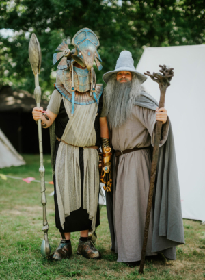 Zwei Teilnehmer des Elbenwald Festivals in fantasievollen Kostümen: der eine verkleidet als ägyptischer Gott mit Speer, der andere als weiser Zauberer mit Stab. Die Veranstaltung kombiniert Musik und Nerd-Kultur in einer lebhaften Festivalatmosphäre.