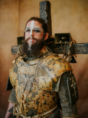 Ein kostümierter Teilnehmer des Elbenwald Festivals trägt eine detaillierte Rüstung aus Papier und Holz, während er vor einem rustikalen Hintergrund posiert. Sein Gesicht ist mit auffälligem Make-up geschminkt, das seine Charakterdarstellung unterstreicht.