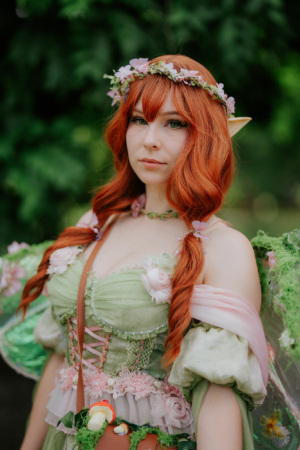 Frau in fantasievoller Kostümierung mit roten Haaren und Blumenkranz, präsentiert sich auf dem Elbenwald Festival. Ihr Outfit besteht aus grünen und rosa Elementen, umgeben von einer natürlichen Kulisse, die die kreative Atmosphäre des Festivals unterstreicht.