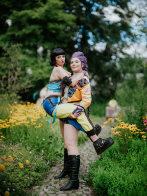 Zwei Cosplayerinnen posieren fröhlich auf einem blühenden Weg beim Elbenwald Festival. Die eine trägt ein farbenfrohes Kostüm mit lila Haaren, während die andere in einem bunten Outfit mit schwarzen Stiefeln lächelt. Ein lebendiger Moment inmitten von Blumen.