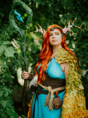 Eine cosplaying Frau mit rotem Haar, die einen bunten Umhang aus Blättern und eine detaillierte Staff hält, steht vor einem grünen Hintergrund. Ihr Outfit ist inspiriert von Fantasy-Elementen und passt perfekt zum Elbenwald Festival.