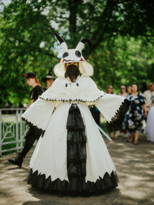Eine kostümierte Teilnehmerin des Elbenwald Festivals trägt ein auffälliges weißes und schwarzes Outfit mit einem großen Kopfteil in Form eines niedlichen, furchterregenden Charakters. Im Hintergrund sind andere Festivalbesucher in bunten Outfits zu sehen.
