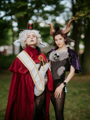 Zwei Cosplayer in aufwendigen Kostümen stehen im Freien beim Elbenwald Festival. Die erste Person trägt ein rotes Cape und eine kreative Kopfbedeckung, während die zweite Person ein tierinspiriertes Outfit mit Hörnern und einem Umhang trägt.