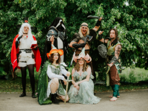 Gruppe von Cosplayern in kreativen Kostümen beim Elbenwald Festival, umgeben von grüner Natur. Die Charaktere zeigen eine Mischung aus Fantasy-Elementen und Musikfestivalkultur, während sie posieren und Spaß haben.