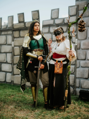 Zwei Frauen in fantasievollen Kostümen stehen vor einer Burgruine auf dem Elbenwald Festival. Die erste trägt eine Rüstung und hält ein Schwert, während die zweite mit einem Stock und einem mystischen Look beeindruckt. Ein kreatives Beispiel für Cosplay und Festivalatmosphäre.