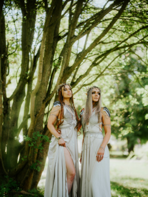 Zwei Frauen in eleganten, fantasievollen Kostümen stehen unter einem Baum beim Elbenwald Festival. Ihre Outfits sind mit glänzenden Elementen verziert, während sie in die Kamera blicken. Die Umgebung strahlt eine magische Atmosphäre aus, die zur Veranstaltung passt.