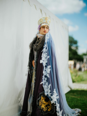 Eine elegant gekleidete Person in einem beeindruckenden Kostüm mit einer kunstvollen Krone und einem langen, verzierten Umhang beim Elbenwald Festival, umgeben von einer grünen Wiese und einem Zelt im Hintergrund, das die festliche Atmosphäre unterstreicht.