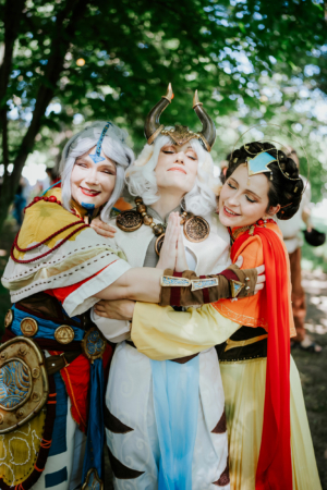 Drei Cosplayerinnen in fantasievollen Kostümen umarmen sich herzlich auf dem Elbenwald Festival, umgeben von grüner Natur. Die Szene strahlt Freude und Gemeinschaft aus, während die Teilnehmer in ihre Rollen schlüpfen und die kreative Atmosphäre genießen.
