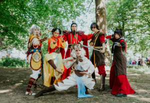 Cosplay-Gruppe beim Elbenwald Festival, gekleidet in aufwendige Kostüme, inspiriert von fantastischen Charakteren. Die Szene spielt im Freien, umgeben von Bäumen und festlicher Atmosphäre, während die Teilnehmer verschiedene Posen einnehmen.