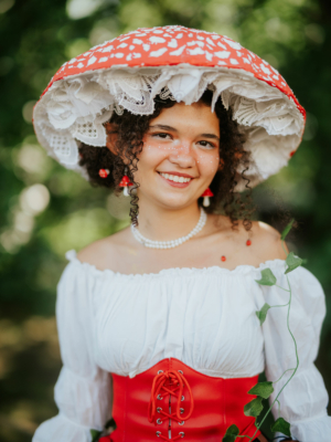 Junge Frau mit einem auffälligen roten Pilzhut und einem weißen Oberteil, die auf dem Elbenwald Festival lächelt. Ihre Frisur ist lockig, und sie trägt ein rotes Korsett sowie eine Perlenkette. Die Umgebung ist grün und sommerlich.