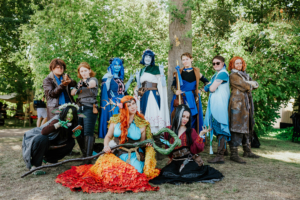 Gruppenselfie von Cosplayern in fantasievollen Kostümen beim Elbenwald Festival, umgeben von grüner Natur. Die Charaktere zeigen eine Vielzahl von Stilen, von magischen Wesen bis zu Abenteurern, und verkörpern die kreative Vielfalt der Veranstaltung.
