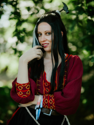 Frau in traditioneller, rot-goldener Kleidung mit langen schwarzen Haaren und einem Hammer im Haar, posiert selbstbewusst im Freien, umgeben von grünen Blättern. Ideal für das Elbenwald Festival, das Musik und Nerd-Kultur vereint.