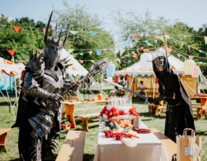 Zwei kostümierte Figuren in beeindruckenden Rüstungen stehen an einem festlich gedeckten Tisch auf dem Elbenwald Festival. Bunte Wimpel und Zelte im Hintergrund schaffen eine lebendige Atmosphäre, während sie mit Requisiten interagieren.