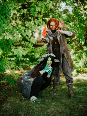 Zwei Cosplayer auf dem Elbenwald Festival posieren vor grüner Kulisse. Der erste trägt eine braune Jacke mit einem feurigen Effekt in der Hand, während der zweite mit grüner Gesichtsbemalung und spitzen Ohren einen Becher hält. Beide verkörpern fantasievolle Charaktere.