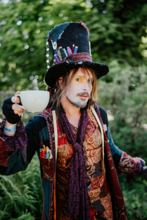 Ein kreativ gekleideter Mann mit einem hohen Hut, der eine Tasse hält. Sein Outfit ist farbenfroh und nostalgisch, geschmückt mit Nähgarn und Accessoires. Die Szene spielt im Freien, umgeben von grüner Natur, passend zur Atmosphäre des Elbenwald Festivals.