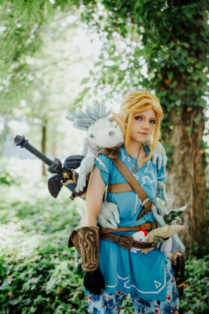 Cosplayer in Link-Kostüm auf dem Elbenwald Festival, umgeben von grüner Natur. Die Person trägt eine Spielzeugwaffe und hat verschiedene Plüschtiere auf dem Rücken. Perfekte Darstellung der Verbindung von Musik und Nerdkultur.