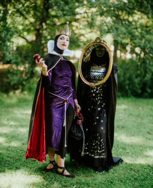 Zwei kostümierte Frauen auf dem Elbenwald Festival: eine in einem lila Kleid mit Krone und Apfel, die andere in schwarzem Gewand mit Spiegel. Die Szenerie zeigt die kreative und fantasievolle Atmosphäre des Festivals im Freien.