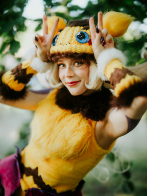 Cosplayer mit buntem Kostüm und tierischen Ohren, die fröhlich Peace-Zeichen macht, vor einer grünen Kulisse auf dem Elbenwald Festival. Das Outfit kombiniert Elemente aus Fantasy und Popkultur, ideal für die kreative Atmosphäre des Events.