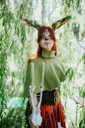 Ein cosplaying Teilnehmer beim Elbenwald Festival, gekleidet in ein grünes Cape und mit kunstvollem Make-up, steht umgeben von hängenden Pflanzen. Sie hält zwei grüne Kerzen in einer dekorativen Halterung und strahlt eine mystische Atmosphäre aus.