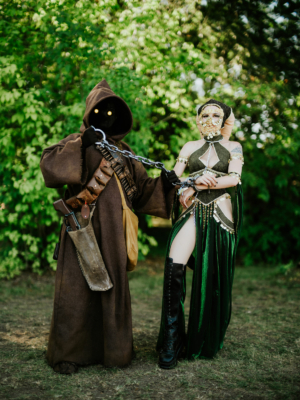 Zwei Cosplayer auf dem Elbenwald Festival: Ein geheimnisvoller Charakter in braunem Gewand mit leuchtenden Augen hält eine Kette, an der eine elegant gekleidete Figur mit goldenen Akzenten und grünem Kleid steht. Hintergrund mit grünen Bäumen.